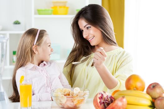 Beautiful young woman gives to the little girl a fruit salad in the kitchen.