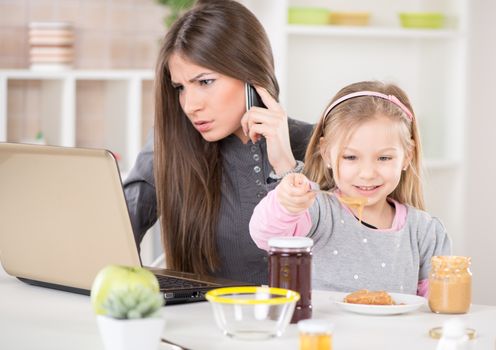 Overworked Business Woman and her little daughter in the morning. Mother read mail and make phone calls before going to work. Daughter smearing peanut butter on bread.