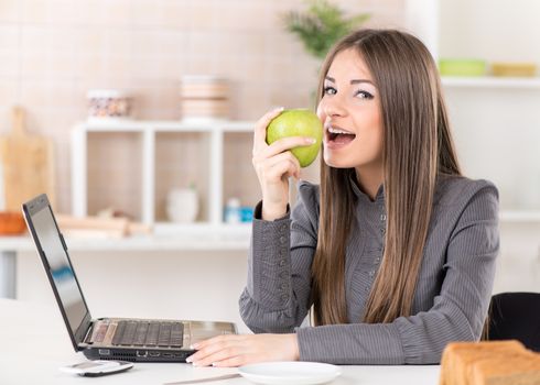 Businesswoman in the kitchen eating apple and reading mail on laptop before going to work.
