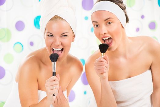 Two young cute woman looking at mirror and preparing to start their day. They having fun and sing with make-up brush.