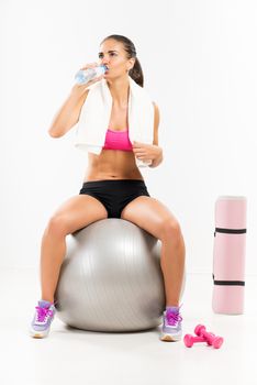 Fitness woman sitting to pilates ball with towel neck and drinking water.