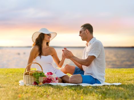 love, dating, people, proposal and holidays concept - smiling young man giving small red gift box with wedding ring to his girlfriend on picnic over seaside sunset background