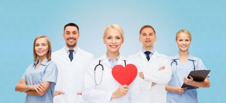 medicine, profession, teamwork and healthcare concept - group of smiling medics or doctors holding red paper heart shape, clipboard and stethoscopes over blue background