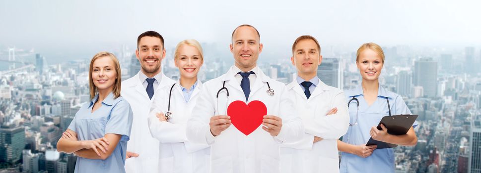 medicine, profession, teamwork and healthcare concept - group of smiling medics or doctors holding red paper heart shape, clipboard and stethoscopes over city background