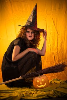 Young woman dressed like a witch. She is in dark clothing with broom and pumpkin.