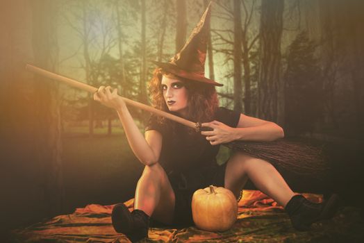 Young woman dressed like a witch. She is sitting in dark clothing with broom and pumpkin.