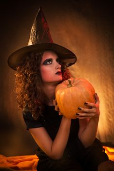 Portrait of Young woman dressed like a witch. She wears dark clothing and holding a pumpkin in hands.