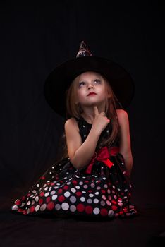 Little halloween witch in dark clothing sitting on black background and thinks.