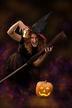 Young woman dressed like a witch. She is in dark with broom and pumpkin.