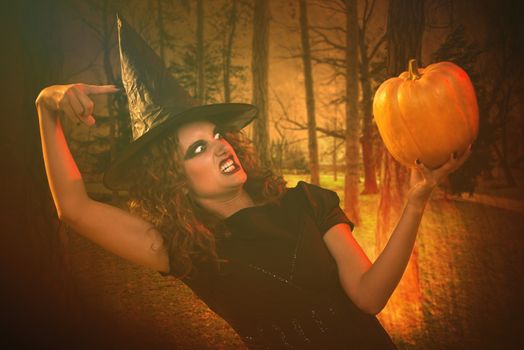 Young woman with evil face dressed like a witch. She wears dark clothing and holding a pumpkin in hand.