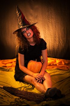 Young woman dressed like a witch. She is in dark and sitting with broom and pumpkin.