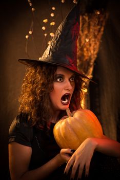 Portrait of young woman with scared face dressed like a witch. She wears dark clothing and holding a pumpkin in hands.