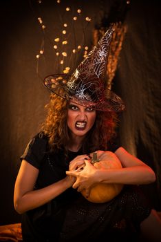 Young woman with evil face dressed like a witch. She wears dark clothing and holding a pumpkin in hands.