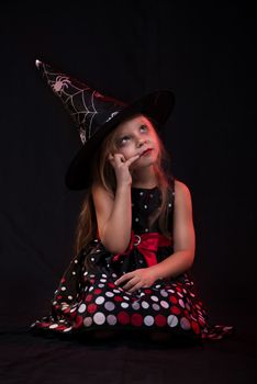 Little halloween witch in dark clothing sitting on black background and thinks.