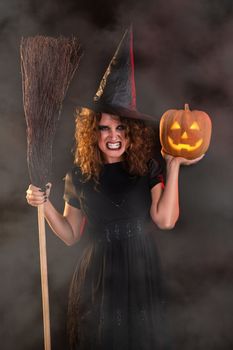 Young woman dressed like a witch. She is in dark clothing and holding broom and pumpkin.