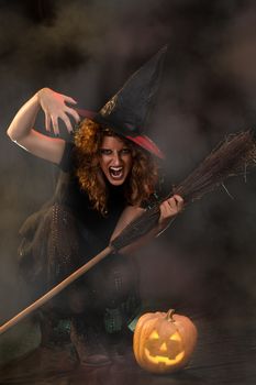 Young woman dressed like a witch. She is in dark with broom and pumpkin.