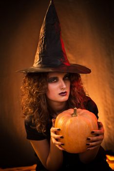 Portrait of Young woman dressed like a witch. She wears dark clothing and holding a pumpkin in hands.