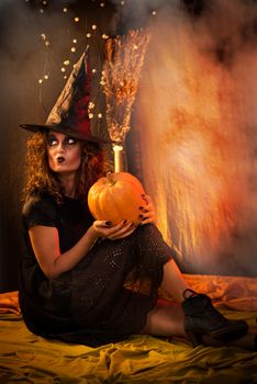 Young woman dressed like a witch. She wears dark clothing and holding a pumpkin in hands.
