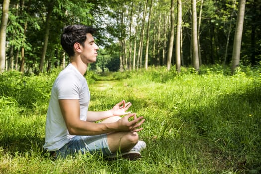 Profile of handsome Young Man During Meditation or Doing an Outdoor Yoga Exercise Sitting Cross Legged on Grassy Ground Alone in Woods