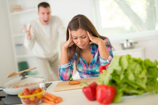 Young couple arguing in the domestic kitchen. Upset woman sitting and holding head. Angry man shouting and waving arms.