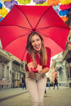Cheerful beautiful young woman with red umbrella in the street decorated with lots of multicolor umbrellas. Looking at camera.
