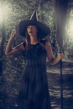 Young woman dressed like a witch at forest. She is in dark clothing and holding a broom. Looking Away.