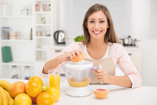 Beautiful young woman in the kitchen squeezing orange juice with an electric juicer. Looking at camera.