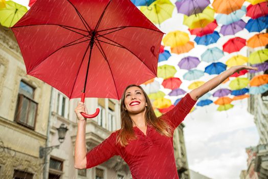 Beautiful young woman walking on the rainy day down the street decorated with lots of multicolor umbrellas. She is holding red umbrella.