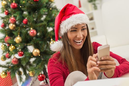 Cheerful young woman wearing Santa's hat and typing message on smart phone at Christmas Time.