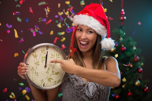 Cheerful beautiful woman celebrating New Year and showing midnight on the clock. She is having fun, confetti is in the air.