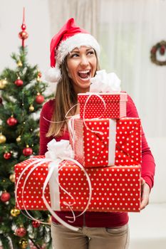 Cheerful beautiful young woman wearing santa hat and holding many Christmas gifts at home.