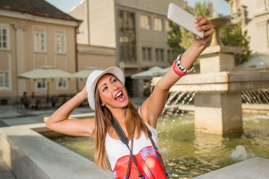 Beautiful young cheerful woman on vacation taking selfie with her smart phone in the city square next to the fountain.