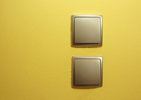 Two flat silver power switch, one below the other on the yellow smooth wall in the room.Close , horizontal view.