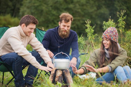 adventure, travel, tourism and people concept - group of smiling friends cooking food in dixie and warming hands sitting around bonfire outdoors