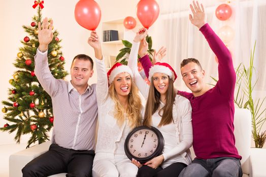 Happy friends Celebrating New Year in home interior with arms raised and showing midnight on the clock.