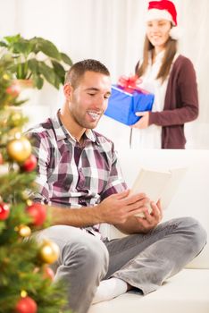 Young beautiful man reading book while his girlfriend holds a Christmas gift and she wants to surprise him.