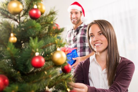 Young beautiful girl decorates the Christmas tree while her boyfriend holds a Christmas gift.