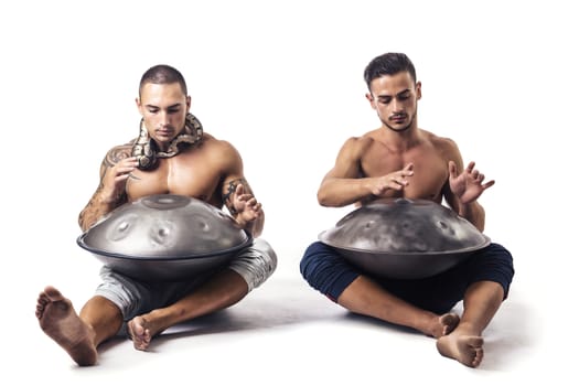 Two Exotic Male Drummers Drumming with Hands on Steel Pan Drums While Seated Beside Each Other in Studio with White Background