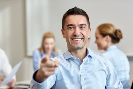 business, people, gesture and teamwork concept - smiling businessman pointing finger to you with group of businesspeople meeting in office