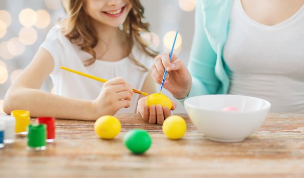 easter, family, holiday, people and childhood concept - close up of happy girl and mother with brushes coloring easter eggs over lights background