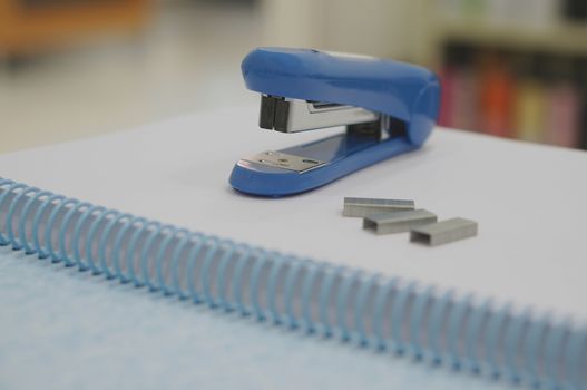Blue stapler, three staples and book placed on table at office.                               