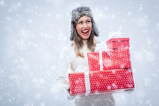 Cheerful beautiful young woman in warm clothing holding many Christmas gifts while snowing.