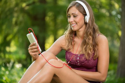 Happy beautiful girl sitting on grass in the park and listening music from smart phone.
