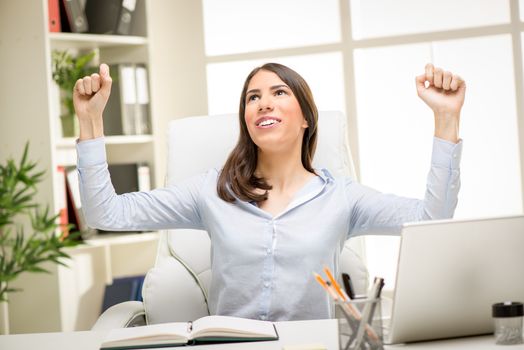Successful businesswoman sitting at the desk in the office with raised arms and celebrating success.