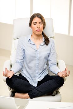 Beautiful young businesswoman practicing yoga in the office. She sits on lotus pose in a chair in front of her desk.
