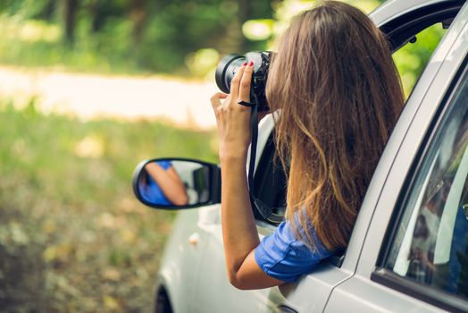 Young beautiful woman sitting in car in the forest and photographing with digital camera.