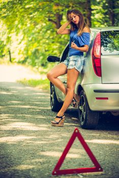 Beautiful young woman with a silver car that broke down on the road in forest. She has set up a warning triangle. Selective focus. Focus on background.