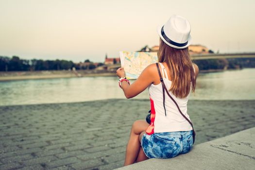 Beautiful young woman on vacation sitting by the river and studying map and looking next direction. Rear view.