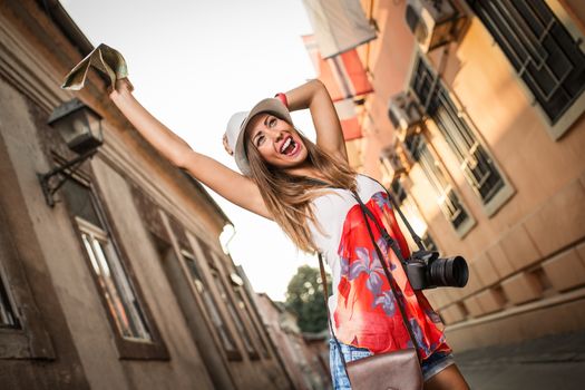 Cheerful woman on vacation walking a narrow street, holding a digital camera and map of city.