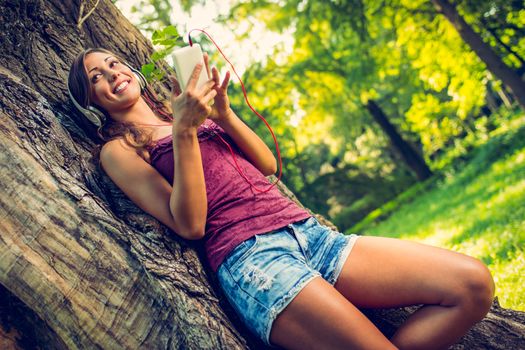 Happy beautiful girl by a tree in a park and listening music from smart phone. 